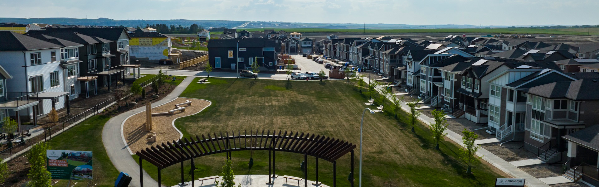 A wide grassy green space with walking paths, wooden seating areas, and a small playground feature, surrounded by rows of modern two-story homes at Ambleton.