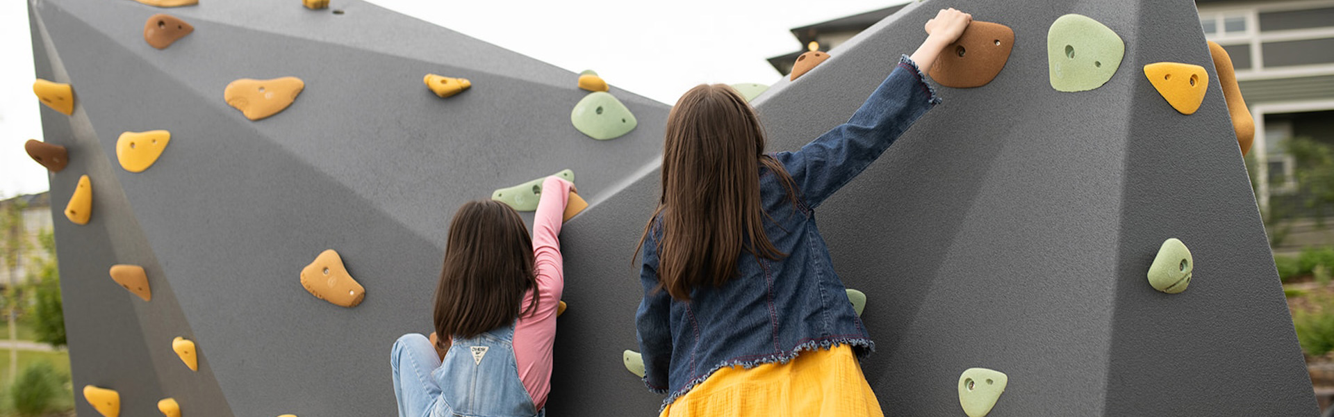 Two girls climbing a grey geometric outdoor rock wall with colourful handholds in a neighbourhood park.