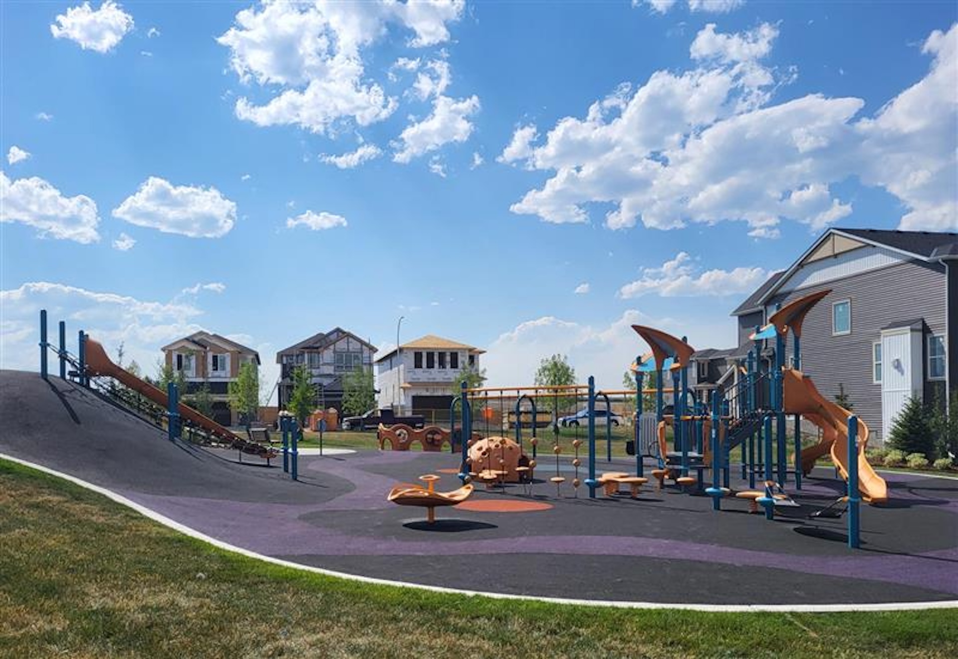Ambleton playground with slides, climbing features, and play equipment set on a rubber surface, surrounded by new residential houses under a bright blue sky with scattered clouds.